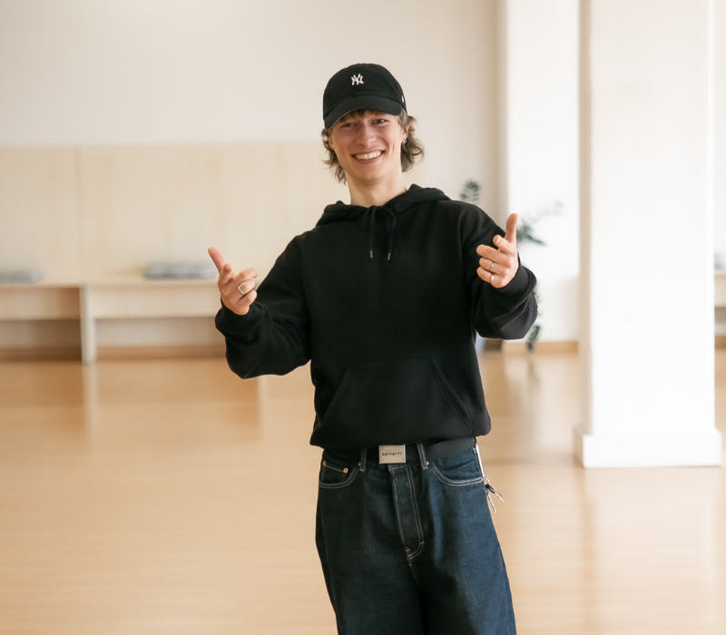 Ein junger Mann steht lächelnd in einem hellen Tanzraum und zeigt mit beiden Händen freundlich auf die Kamera. Er trägt einen schwarzen Hoodie, eine dunkle Jeans und eine schwarze Baseballcap. Im Hintergrund sind helle Wände, Sitzbänke und eine Zimmerpflanze zu sehen.