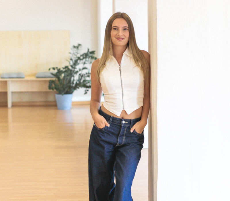 Eine junge Frau mit langen, glatten Haaren lehnt entspannt an einer weißen Säule in einem hellen Tanzraum. Sie trägt ein ärmelloses, weißes Oberteil mit Reißverschluss und eine weite blaue Jeans. Ihre Hände stecken locker in den Hosentaschen und sie lächelt freundlich in die Kamera. Im Hintergrund stehen eine Bank und eine grüne Zimmerpflanze.
