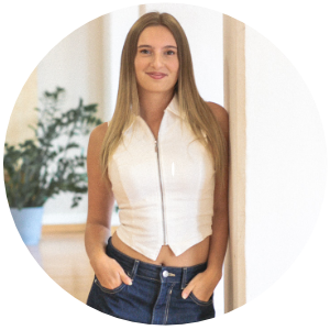 Porträt einer jungen Frau, die entspannt an einer weißen Säule in einem hellen Tanzraum lehnt. Sie trägt ein ärmelloses weißes Oberteil mit Reißverschluss und eine weite Jeans, die Hände locker in den Taschen. Im Hintergrund stehen eine grüne Zimmerpflanze und eine helle Bank.