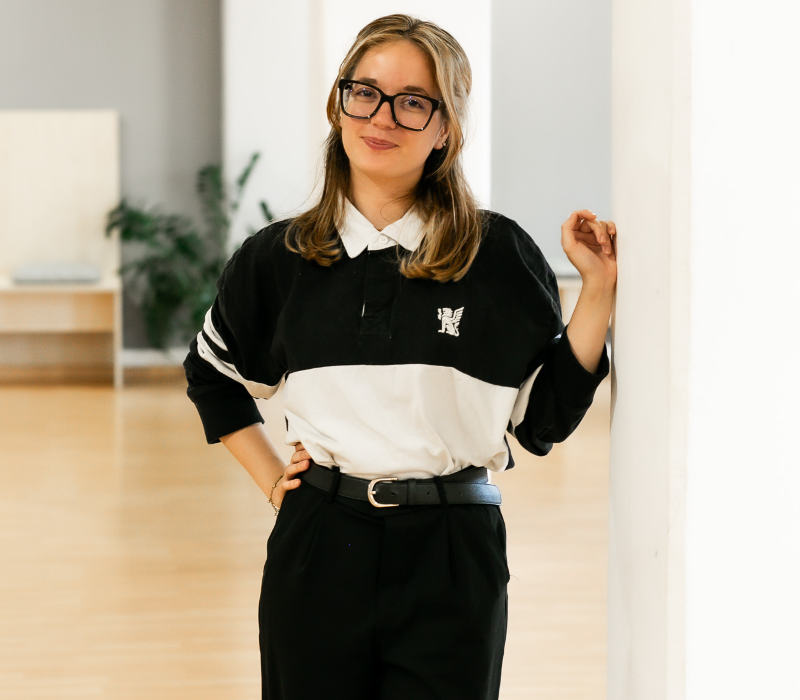 Eine junge Frau mit langen, hellbraunen Haaren und großer Brille steht lässig in einem hellen Tanz- oder Bewegungsraum. Sie trägt ein schwarz-weißes Rugbyshirt mit Logo, eine schwarze Hose und einen Gürtel. Mit einer Hand stützt sie sich an einer weißen Säule ab und lächelt selbstbewusst in die Kamera.